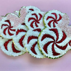 May include: A collection of embroidered patches shaped like mouse ears with a peppermint candy pattern. The patches feature a white and red swirl design on a textured white base, accented with light pink fuzzy fabric and a gold metallic border.