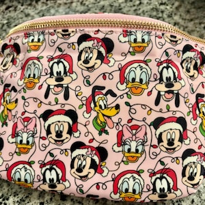 May include: A light pink fanny pack featuring a repeating pattern of Disney characters including Mickey Mouse, Minnie Mouse, Donald Duck, Daisy Duck, Goofy, and Pluto, all wearing Santa hats, interspersed with colorful Christmas light strings and a gold zipper.