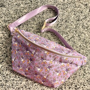 May include: A light purple fanny pack featuring an all-over print of Daisy Duck characters and small yellow flowers. The bag includes an adjustable matching purple strap and a gold-toned metal zipper closure across the top main compartment.