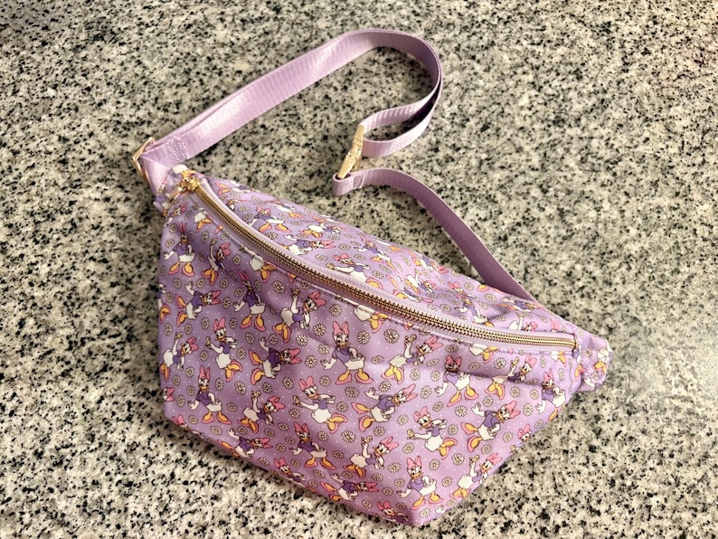May include: A light purple fanny pack featuring an all-over print of Daisy Duck characters and small yellow flowers. The bag includes an adjustable matching purple strap and a gold-toned metal zipper closure across the top main compartment.