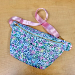 May include: A fanny pack featuring a light blue background with a repeating pattern of pink flowers, purple blossoms, green leaves, seashells, and blue carriage motifs. The bag has a gold-toned metal zipper closure and an adjustable light pink strap.