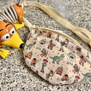 May include: A cream-colored fanny pack featuring a repeating pattern of Toy Story characters including Woody, Buzz Lightyear, Rex, Slinky Dog, and Mr. Potato Head, with a gold zipper and an adjustable strap, positioned next to a Slinky Dog toy.