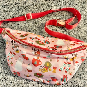 May include: A light pink fanny pack featuring a repeating holiday pattern of Mickey Mouse, Pluto, snowmen, Christmas stockings, wrapped gifts, wreaths, and mugs. The bag has a gold-toned zipper closure and an adjustable bright red nylon strap.
