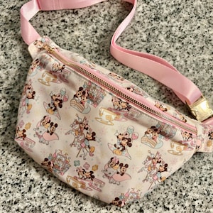 May include: A light pink fanny pack featuring a repeating pattern of Mickey and Minnie Mouse characters, Dumbo, and teacup ride icons. The bag includes a gold-toned metal zipper closure and an adjustable light pink nylon strap with a gold buckle.