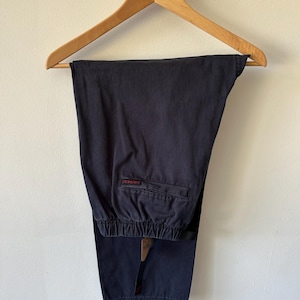 Peut inclure: Un pantalon noir uni doté d'une taille élastique et d'une poche arrière unique avec une petite étiquette rectangulaire rouge portant du texte noir. Le pantalon est fabriqué dans un tissu mat et est présenté suspendu à un cintre en bois.