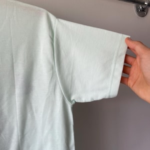 May include: A light mint green short-sleeved t-shirt featuring a simple crew neckline and a plain, solid-colored fabric construction. The garment is shown from a side angle, highlighting the sleeve seam and the smooth texture of the material.