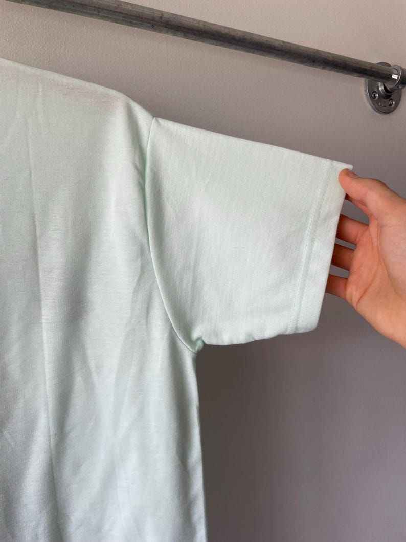 May include: A light mint green short-sleeved t-shirt featuring a simple crew neckline and a plain, solid-colored fabric construction. The garment is shown from a side angle, highlighting the sleeve seam and the smooth texture of the material.