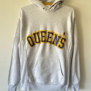 May include: A light grey hooded sweatshirt featuring the word QUEENS in large, arched, blue and yellow embroidered lettering across the chest. The garment includes a front kangaroo pocket, long sleeves with ribbed cuffs, and a drawstring hood.