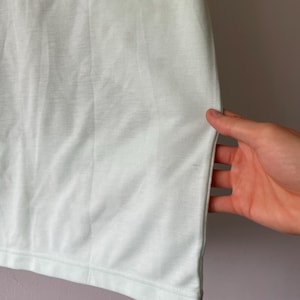 May include: A plain, light mint green t-shirt featuring a smooth, solid-colored fabric with a visible hemline at the bottom edge. The garment is constructed with a simple, clean design and a consistent, soft-toned pastel green color throughout.