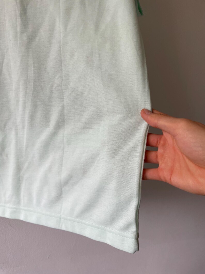 May include: A plain, light mint green t-shirt featuring a smooth, solid-colored fabric with a visible hemline at the bottom edge. The garment is constructed with a simple, clean design and a consistent, soft-toned pastel green color throughout.