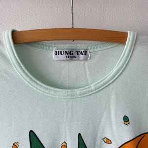 May include: A light mint green t-shirt with a crew neckline and a white label reading HUNG TAT FASHION. The fabric features a graphic print of small orange acorns and a large orange umbrella shape against the solid light green background.