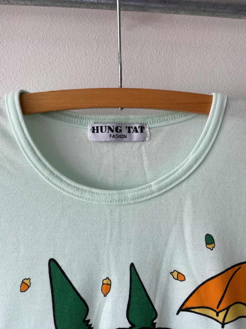 May include: A light mint green t-shirt with a crew neckline and a white label reading HUNG TAT FASHION. The fabric features a graphic print of small orange acorns and a large orange umbrella shape against the solid light green background.
