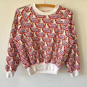 May include: A white long-sleeve sweatshirt featuring an all-over repeating pattern of the Planters Peanuts logo and the Mr. Peanut character. The design includes red triangular shapes with white text and a ribbed white crew neck and cuffs.