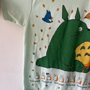 May include: A light mint green t-shirt featuring a large green character with a yellow belly, a small blue character, and a row of tiny white and orange creatures. The text "TOTORO" is printed repeatedly below the small creatures, surrounded by small orange acorns.