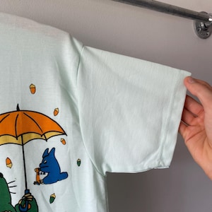 May include: A light mint green short-sleeve t-shirt featuring a graphic print of a large green creature holding a bright orange umbrella, with a smaller blue creature and several scattered brown acorns printed on the front of the fabric.
