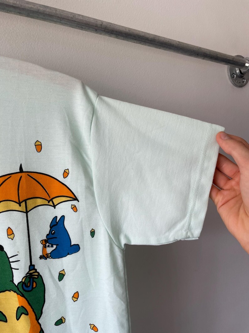 May include: A light mint green short-sleeve t-shirt featuring a graphic print of a large green creature holding a bright orange umbrella, with a smaller blue creature and several scattered brown acorns printed on the front of the fabric.