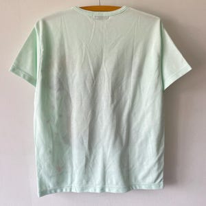 May include: A short-sleeved t-shirt in a pale mint green color. The garment features a crew neckline and is displayed hanging from a wooden hanger. The fabric has a smooth texture with subtle, faint reddish-brown markings visible on the left side.