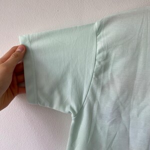 May include: A light mint green short-sleeved t-shirt made of a smooth, solid-colored fabric. The garment features a simple crew neckline and a standard sleeve construction with visible stitching along the hem and shoulder seams of the sleeve.
