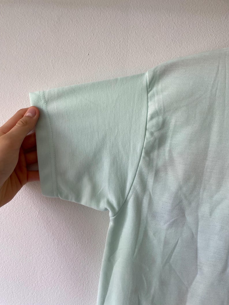May include: A light mint green short-sleeved t-shirt made of a smooth, solid-colored fabric. The garment features a simple crew neckline and a standard sleeve construction with visible stitching along the hem and shoulder seams of the sleeve.