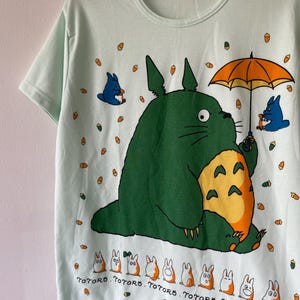May include: A light mint green short-sleeve t-shirt featuring a large green Totoro character holding an orange umbrella, surrounded by small acorns, blue figures, and a row of white and orange creatures with the text Totoro printed underneath.
