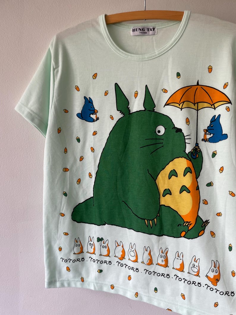 May include: A light mint green short-sleeve t-shirt featuring a large green Totoro character holding an orange umbrella, surrounded by small acorns, blue figures, and a row of white and orange creatures with the text Totoro printed underneath.