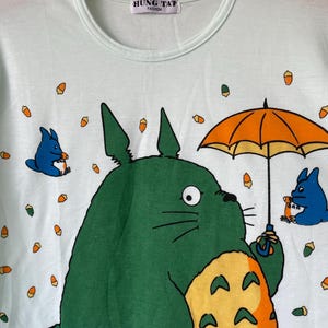 May include: A light mint green t-shirt featuring a large green creature with a yellow belly holding an orange umbrella, accompanied by two smaller blue creatures and scattered orange acorns. The inner neck label reads HUNG TAT FASHION.