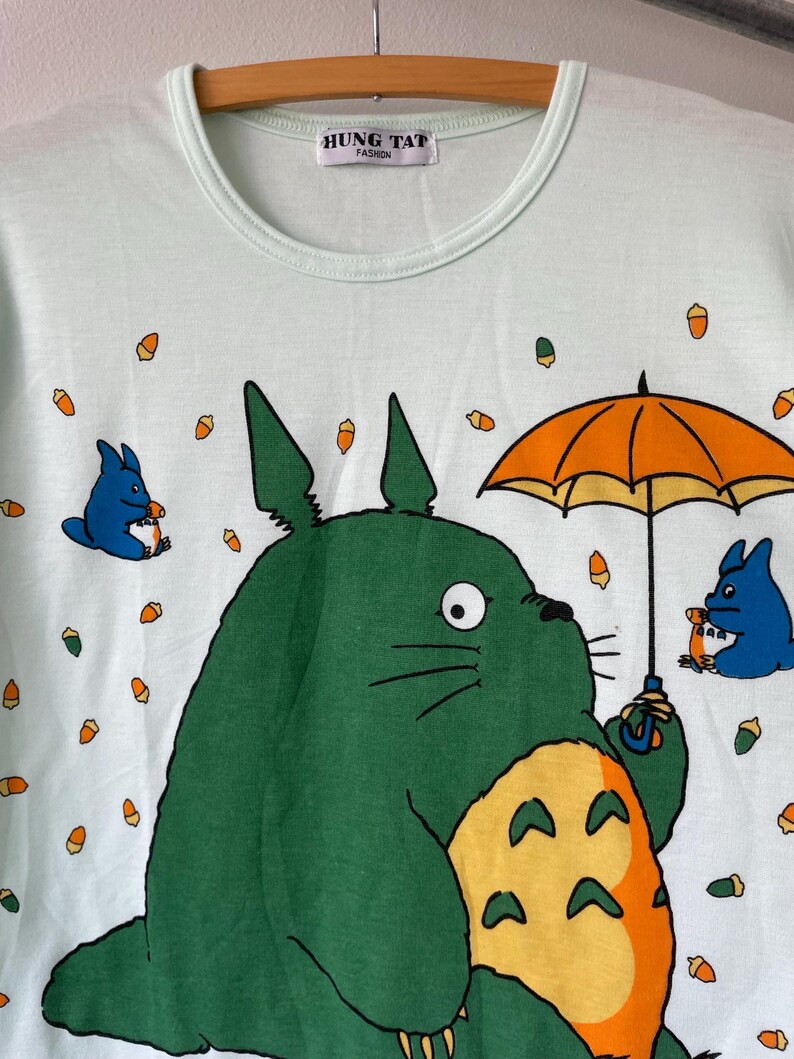 May include: A light mint green t-shirt featuring a large green creature with a yellow belly holding an orange umbrella, accompanied by two smaller blue creatures and scattered orange acorns. The inner neck label reads HUNG TAT FASHION.