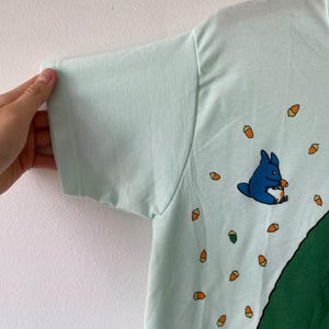 May include: A light mint green t-shirt featuring a small blue creature illustration holding an acorn, surrounded by a pattern of scattered orange and brown acorns, with a large dark green shape visible along the bottom right side of the garment.