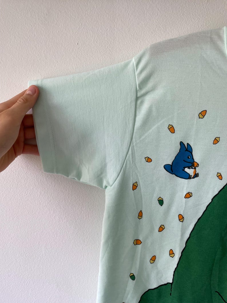 May include: A light mint green t-shirt featuring a small blue creature illustration holding an acorn, surrounded by a pattern of scattered orange and brown acorns, with a large dark green shape visible along the bottom right side of the garment.