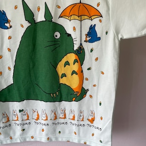 May include: A white t-shirt featuring a large green creature with a yellow belly holding an orange umbrella. Smaller blue creatures and orange acorns are scattered across the fabric. A row of small white and orange figures appears at the bottom with the text TOTORO.