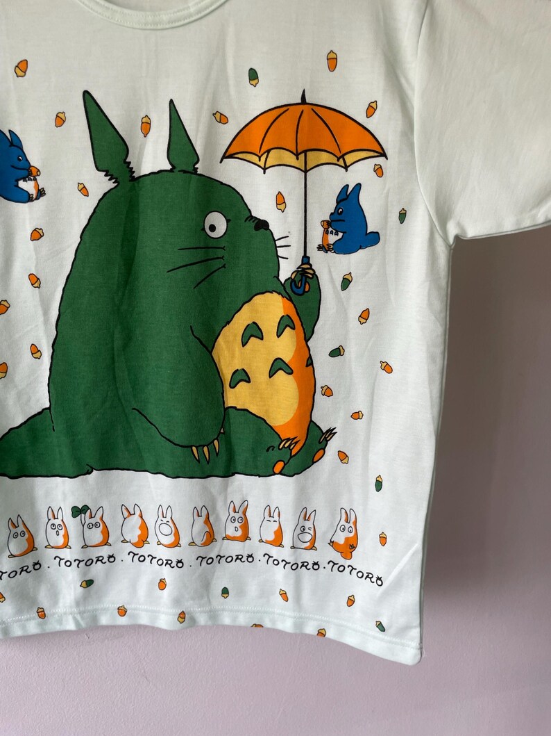 May include: A white t-shirt featuring a large green creature with a yellow belly holding an orange umbrella. Smaller blue creatures and orange acorns are scattered across the fabric. A row of small white and orange figures appears at the bottom with the text TOTORO.