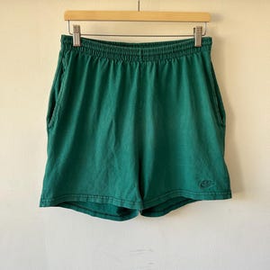 以下が含まれることがあります： ウエストゴムとサイドポケットを備えたフォレストグリーンのアスレチックショーツ。右脚下部には同系色の小さなNikeロゴが刺繍されています。生地はわずかに色あせた使い込まれた風合いで、縫い目がはっきりと見えます。