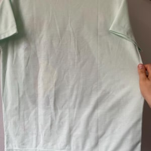 May include: A short-sleeved t-shirt in a pale mint green color. The garment features a crew neckline and a straight hemline. A faint, indistinct graphic print is visible on the front center of the shirt, showing subtle variations in tone.