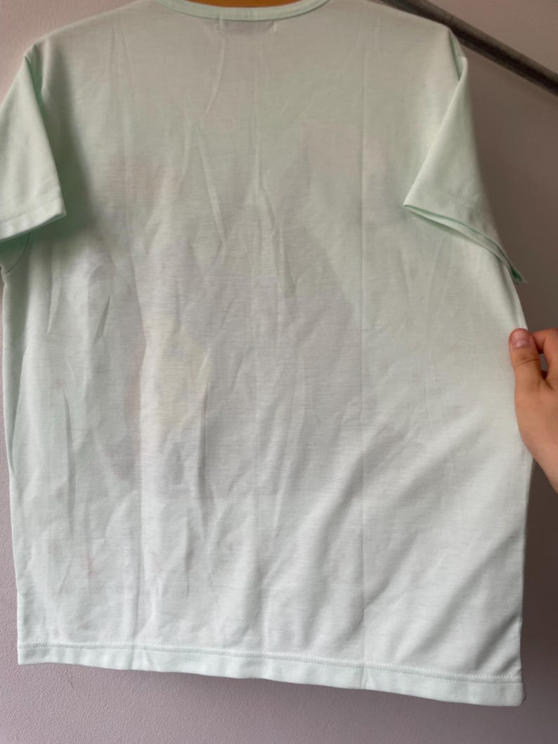 May include: A short-sleeved t-shirt in a pale mint green color. The garment features a crew neckline and a straight hemline. A faint, indistinct graphic print is visible on the front center of the shirt, showing subtle variations in tone.