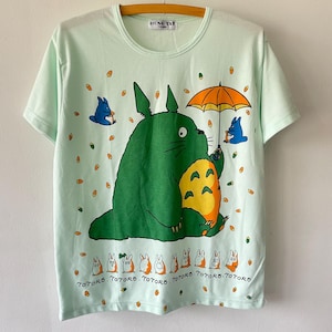May include: A light mint green short-sleeve t-shirt featuring a large green Totoro character holding an orange umbrella, surrounded by small blue creatures and orange acorns. The text "TOTORO" is printed repeatedly below a row of small characters.