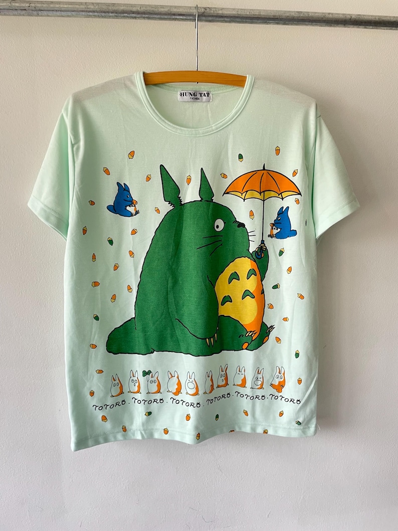 May include: A light mint green short-sleeve t-shirt featuring a large green Totoro character holding an orange umbrella, surrounded by small blue creatures and orange acorns. The text "TOTORO" is printed repeatedly below a row of small characters.