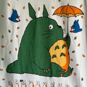 May include: A light green t-shirt featuring a large green Totoro character holding an orange umbrella, surrounded by small blue creatures and orange acorns. At the bottom, a row of small white and orange creatures appears with the text Totoro.
