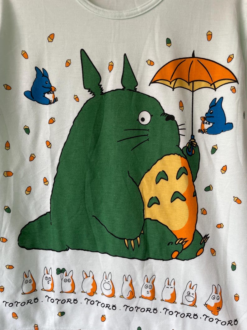 May include: A light green t-shirt featuring a large green Totoro character holding an orange umbrella, surrounded by small blue creatures and orange acorns. At the bottom, a row of small white and orange creatures appears with the text Totoro.