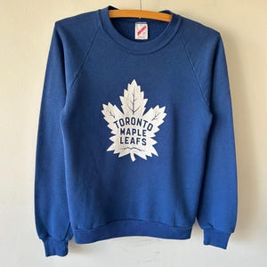 May include: A blue long-sleeved crewneck sweatshirt featuring a large white graphic of a maple leaf on the chest. The text TORONTO MAPLE LEAFS is printed in blue inside the white leaf graphic. The garment has a ribbed collar, cuffs, and hem.