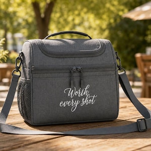 Könnte beinhalten: Eine anthrazitfarbene isolierte Kühltasche mit Tragegriff und verstellbarem Schultergurt. Auf der Vorderseite befindet sich der weiße kursive Schriftzug Worth every shot. Sie verfügt über ein Reißverschluss-Hauptfach und eine seitliche Netztasche.