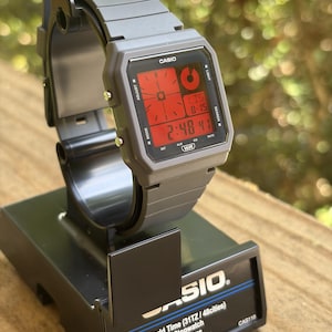May include: A grey Casio digital watch with a red display screen featuring a square case and matching resin strap. The watch face shows the time 2:48 and date 8-15, with text including CASIO, ADJUST, LIGHT, MODE, SEARCH, DST, ALM, SIG, MUTE, and WR.