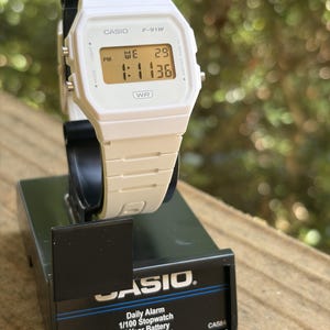 May include: A white Casio F-91W digital watch with a rectangular face, a light-colored LCD screen displaying the time 1:11:36, the day WE, and the date 29. The watch features a white resin strap and is mounted on a black display stand with text.