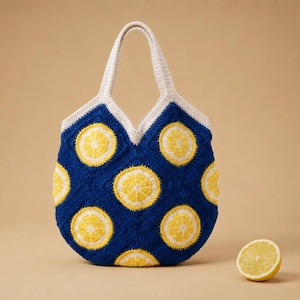 May include: A crochet tote bag featuring a deep blue background with a repeating pattern of yellow and white lemon slice motifs. The bag has two integrated cream-colored handles and a rounded shape. A single fresh lemon half sits to the right.
