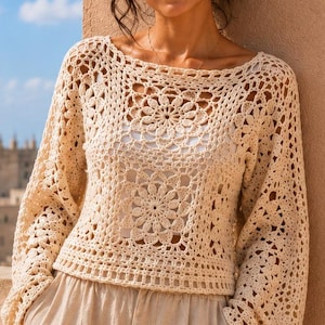 May include: A cream-colored long-sleeve crochet sweater featuring a prominent floral motif pattern, a wide boat neckline, and loose, flared sleeves, paired with light beige trousers, showcasing an intricate openwork knit design throughout.