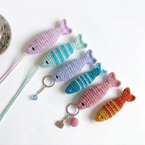 Könnte beinhalten: Eine Sammlung von sechs gehäkelten Fischornamenten in verschiedenen Farben wie Lavendel, Hellblau, Türkis, Rosa und Orange. Einige Fische haben Streifenmuster, andere sind einfarbig. Zwei Fische haben lange Garnschwänze und zwei haben Metallschlüsselanhänger.