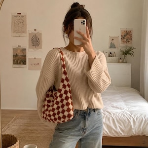 May include: A beige ribbed knit sweater with long sleeves and a crew neckline, paired with light blue denim jeans and a brown and white checkered crochet shoulder bag. The bag features a repeating diamond pattern and a single integrated strap.