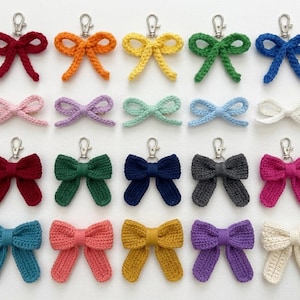 May include: A collection of twenty crocheted bow keychains arranged in four rows. The top row features five bows with silver lobster clasps in red, orange, yellow, green, and blue. The remaining rows display various colored bows in different sizes.