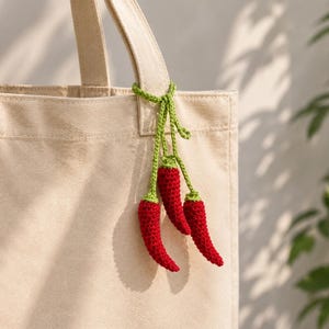Op de afbeelding: Een beige canvas draagtas met een decoratieve gehaakte hanger aan het handvat. De hanger bestaat uit drie kleine, rode chilipepers met groene steeltjes, verbonden door dunne groene gehaakte koordjes die aan de tasriem naar beneden hangen.