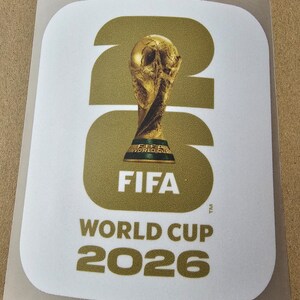 Puede incluir: Una pegatina rectangular con esquinas redondeadas que presenta el trofeo dorado de la Copa Mundial de la FIFA centrado sobre los grandes números dorados 26. Debajo del trofeo, el texto dice FIFA, y en la parte inferior, WORLD CUP 2026 en letras doradas.