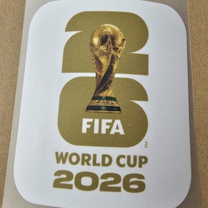 Può includere: Un adesivo rettangolare con angoli arrotondati che presenta il numero 26 di colore oro sullo sfondo. Al centro si trova il trofeo della Coppa del Mondo FIFA sopra il testo bianco FIFA e il testo dorato WORLD CUP 2026 su fondo bianco.